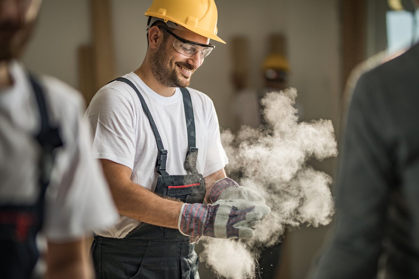 Happy construction worker cleaning his gloves from sawdust at construction site.